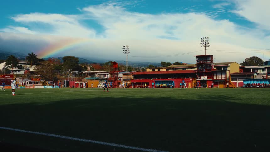 Players in blue and white jerseys compete for the ball on a lush green soccer field while a referee with a flag moves into view all set against the backdrop of a mountain stadium and a brilliant rainbow stretching across the sky