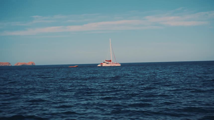 A white catamaran sailboat floats on the blue ocean with rocky islands in the distance