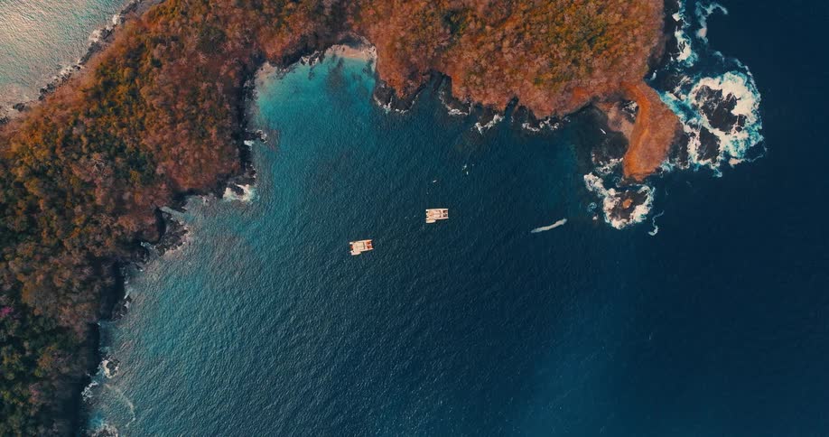 Two yachts float peacefully in a deep blue bay as the perspective lowers toward the water revealing a jagged coastline covered in dense autumn foliage and crashing white sea foam