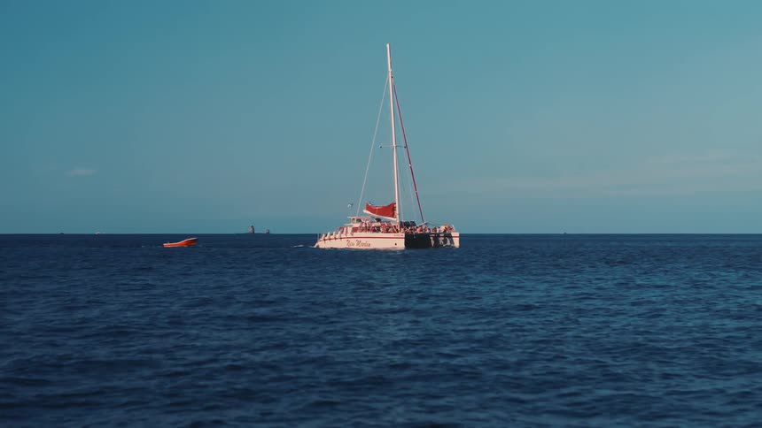 A large catamaran sailboat filled with people floats on the open blue ocean under a clear sky