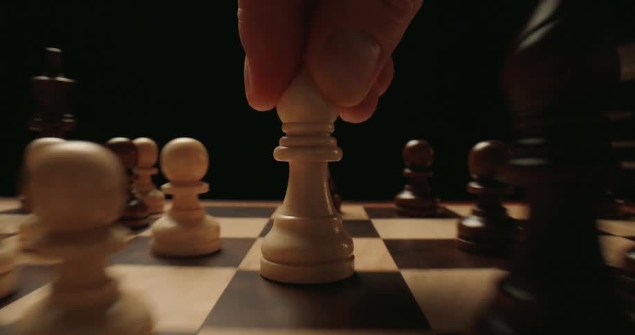 A close view shows a person moving a white pawn across a wooden chess board during a game against a dark background