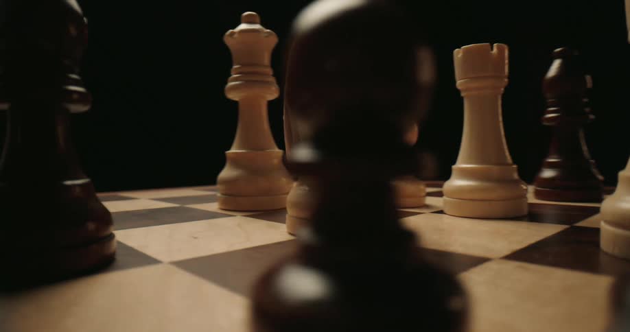 Wooden chess pieces are positioned on a chessboard under low lighting