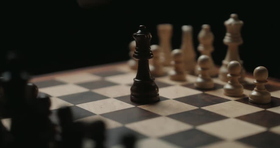 The camera orbits a wooden chess board as a hand reaches into the frame to place a black piece capturing a white piece and completing a strategic move within the moody lowlight atmosphere of the quiet room