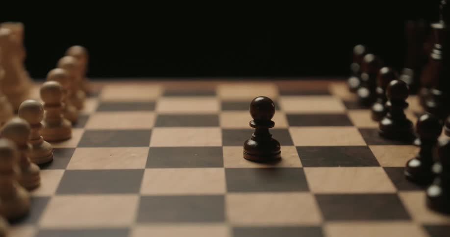 A close view shows a person moving a black pawn forward on a wooden chess board to start a game against a dark background