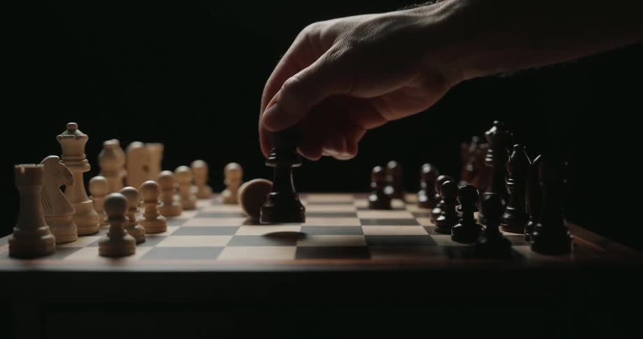 A close view shows a person using a black king to knock over a white king on a wooden chess board during a game against a dark background