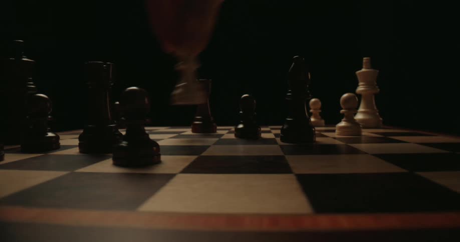 A close view shows a person moving a black bishop and other pieces across a wooden chess board during a game against a dark background
