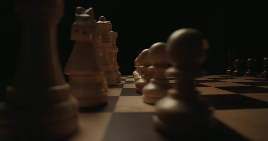 A close view of a chess board shows white wooden pieces arranged on brown and tan squares against a dark background in a dim room