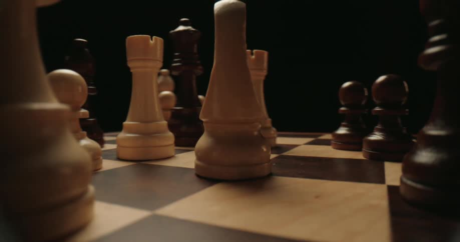 The camera glides at a low angle across a wooden chessboard weaving through cream and dark brown chess pieces that stand in a strategic formation against a dark moody background