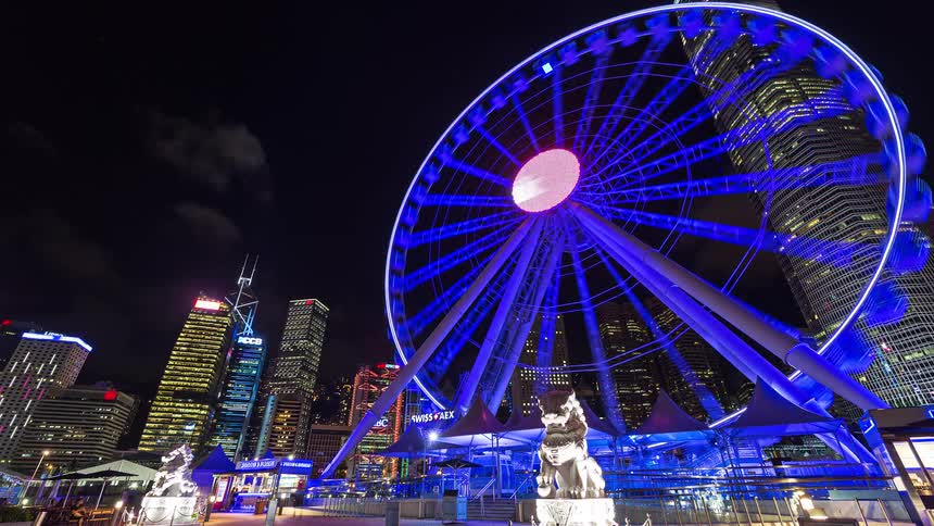 Glowing lights on downtown buildings and towers overlook a ferris wheel turning as people walk through the midway
