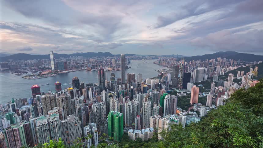 A wide view from a high vantage point shows a dense cityscape of tall skyscrapers and residential buildings along a harbor as day turns to night and city lights begin to glow