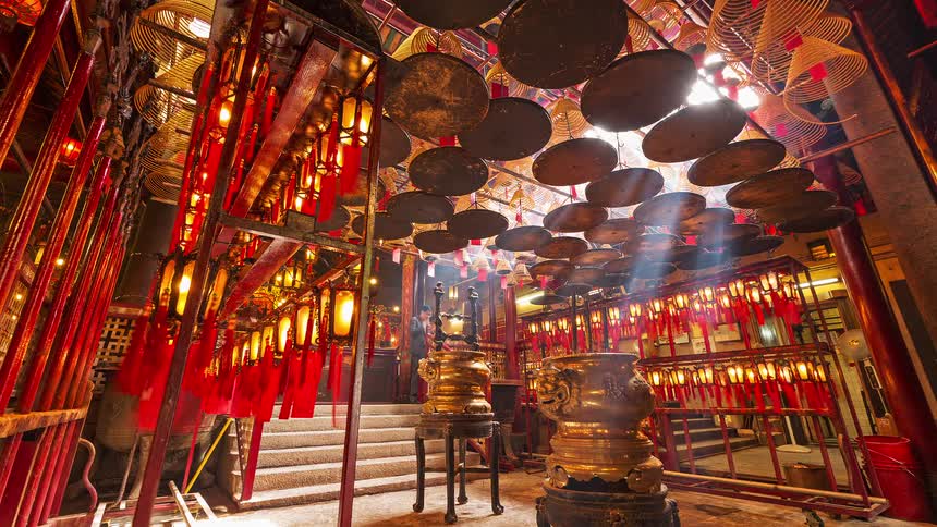 A view inside a traditional temple shows many circular incense coils and red lanterns hanging from the ceiling above golden incense burners and stone steps in a warm and dimly lit atmosphere