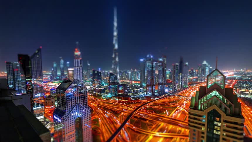 A high angle time lapse view at night shows the illuminated skyscrapers of dubai with the burj khalifa standing tall over glowing orange light trails from traffic on a large highway interchange
