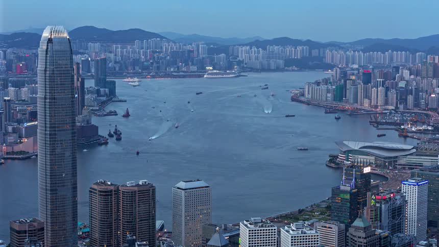 A high angle timelapse shows a city as day turns to night featuring heavy boat traffic while city lights begin to flicker on against the darkening blue sky and distant hills