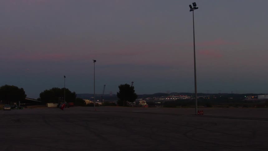 A vast empty asphalt lot under a deep purple and pink twilight sky where tall light poles stand tall against a distant industrial horizon featuring silhouettes of trees and structural cranes in the fading evening light