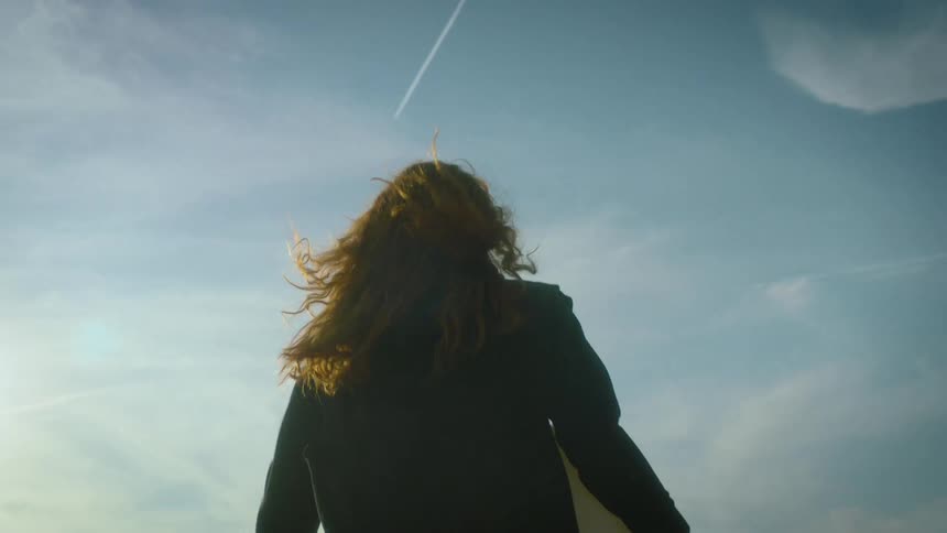 The video shows a woman with long reddish hair running forward while holding a white objecther hair blows wildly in the wind against a bright clear sky as she moves