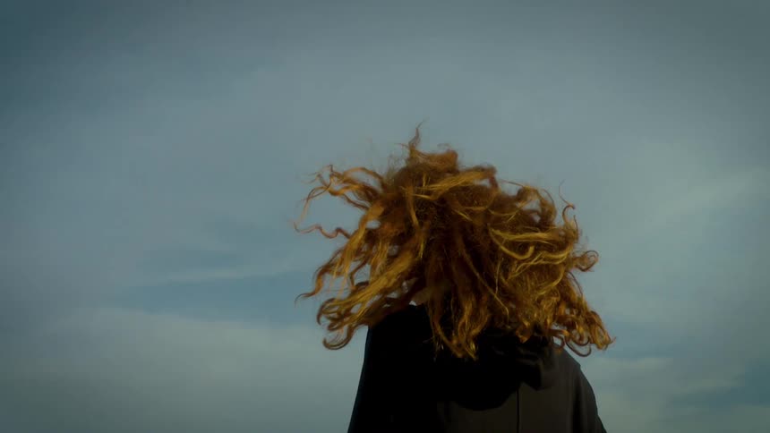 A person with long ginger hair stands outside against a cloudy sky their hair blowing dramatically in the windthe camera captures them from a low angle as they turn away focusing on the movement of their hair and the muted cinematic li
