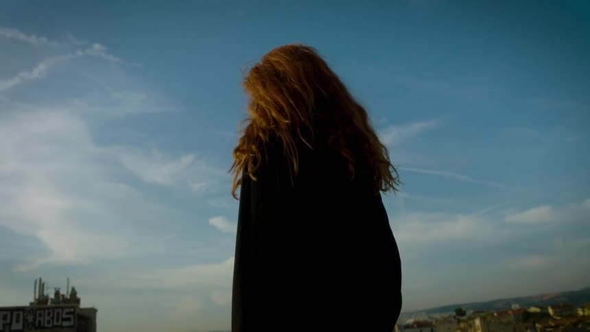 A woman with long red hair stands atop a building overlooking a city