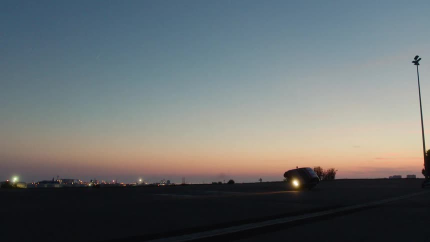 A small three wheeled vehicle drives along a paved road at dusk with city lights in the distance