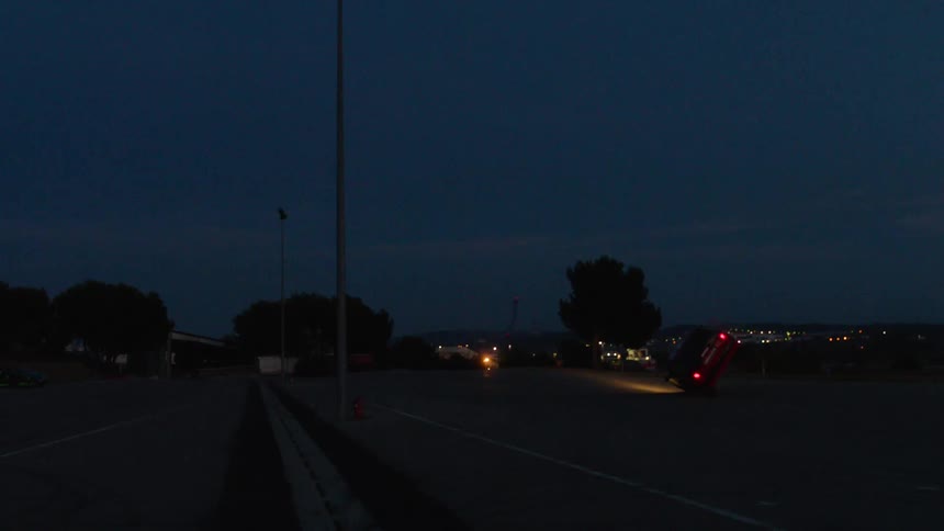 A car tipping onto two wheels while driving across a large paved area at night with city lights visible in the background