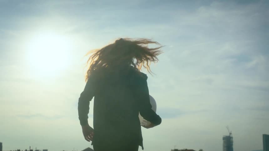 A woman with long curly hair silhouetted against a bright sky walks while holding a white object