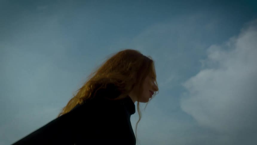A view from below shows a woman with long red hair wearing black clothing as she looks around and runs against a blue sky with white clouds