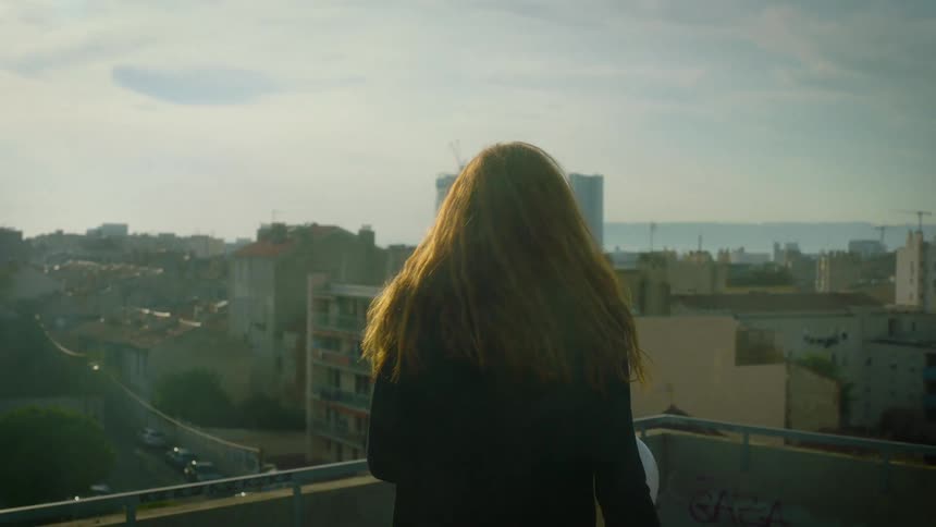 A woman with long reddish hair stands on a rooftop overlooking a city as the wind blows through her hairthe camera remains behind her capturing the vast urban landscape and the hazy horizon under a bright sky