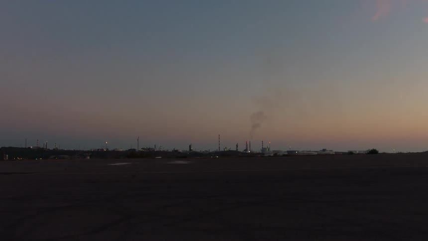 As dusk settles a sprawling industrial complex with tall smokestacks stands silhouetted against a fading purple and orange horizon with faint plumes of smoke drifting into the darkening sky