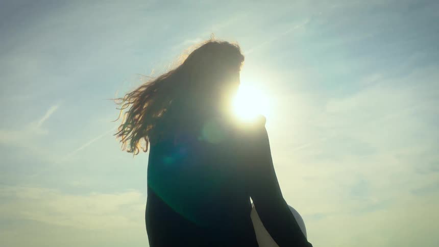 A young woman walking outdoors under the orange sunset holding a white hard hat as her long hair swings in the wind