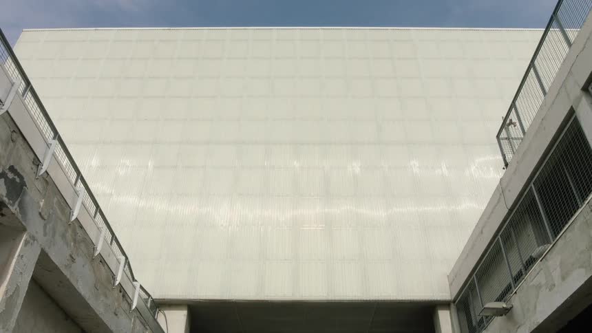 A view looking up from between two concrete structures shows the smooth white facade of a large building against a clear blue sky