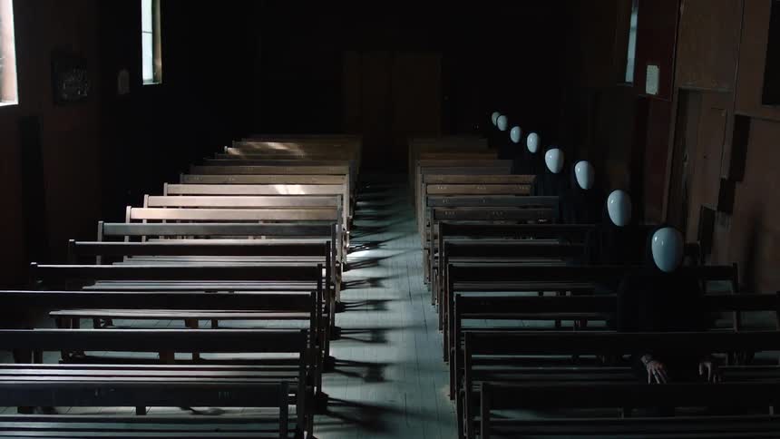 A view inside a dimly lit room shows rows of wooden benches with several figures wearing black clothing and white masks sitting still in the shadows