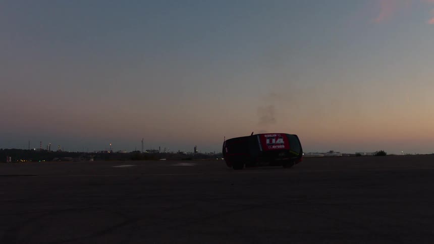 A black and red racing car numbered 114 performs a stunt by driving on two wheels across a dark open field at dusk with a large industrial oil refinery and glowing factory lights visible along the horizon under a clear twilight sky