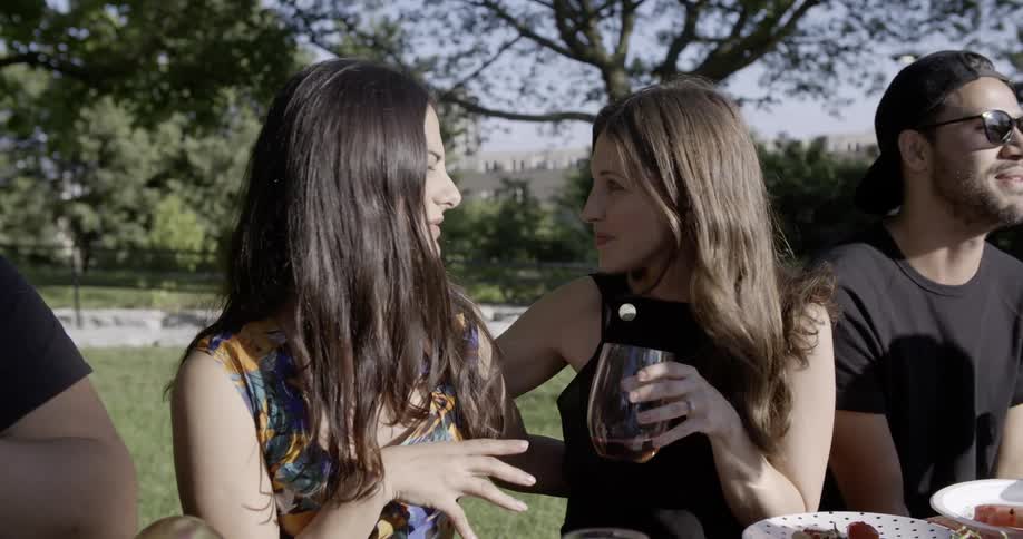 Two women are talking at a picnic table with other people on sunny day