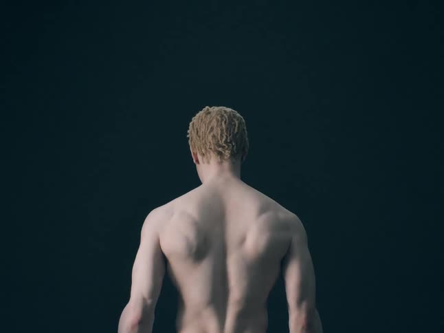 A view from behind shows the muscular back and shoulders of a shirtless man with short blonde hair standing still against a solid dark background in a dim studio