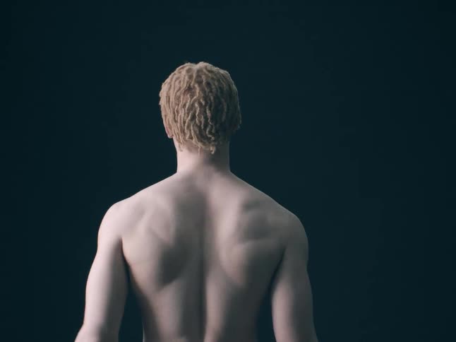 A view from behind shows the muscular back and shoulders of a man with short blonde hair gesturing with his arms against a solid dark background in a dim studio