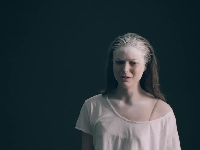 A young woman stands against a green wall with a white substance on her face looking frightened