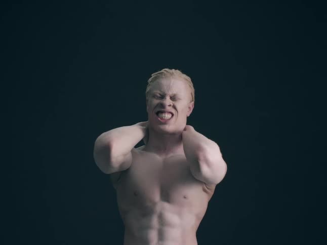 A shirtless man with blonde hair and pale skin clinches his jaw and holds the back of his neck with both hands against a solid dark background in a dim studio