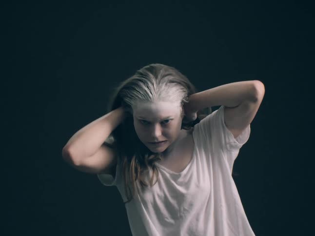 The video shows a woman with white paint or powder covering the top of her head and forehead wearing an oversized white tshirt she holds her hands behind her head and stares intensely at the camera with a strained emotional expression against a dark b