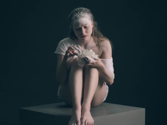 A woman with pale skin and light hair sits curled on a dark pedestal holding a delicate white flowershe appears to be in a moment of quiet contemplation as she looks down at the petals in a dimly lit minimalist setting