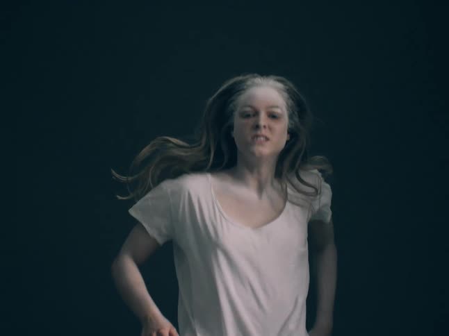 The video shows a woman with pale skin and lightcolored hair running toward the camera against a dark minimalist backgroundher expression is intense and focused while her hair and white tshirt flow behind her to emphasize the sense of motion