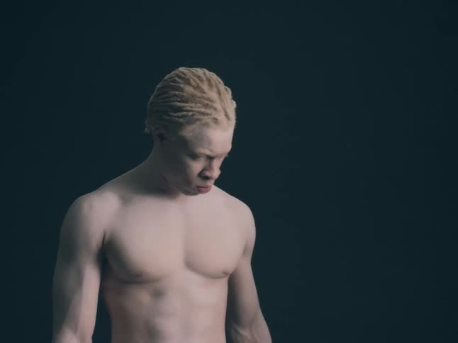 A close view shows a person with a pale complexion and short blonde hair looking down while standing shirtless against a dark background