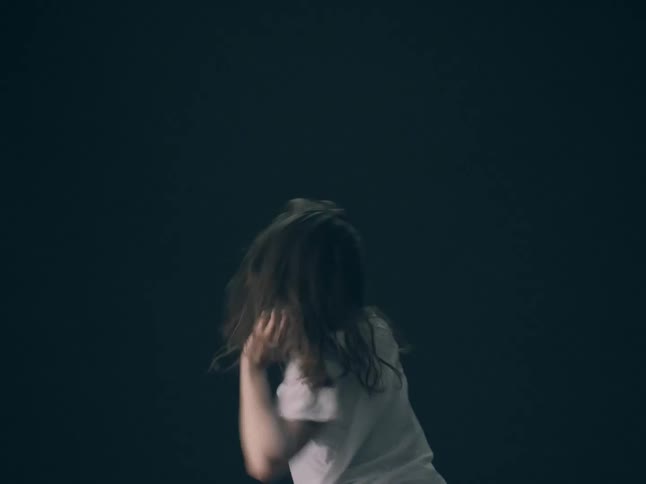 A close view shows a person with long hair and white powder on their face as they move their head and arms aggressively against a dark background