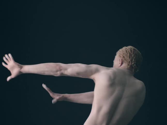 A man with blonde textured hair performs a fluid expressive dance movement against a dark minimalist backgroundhis muscles are highlighted by soft lighting as he extends his arms outward in a graceful sweeping motion