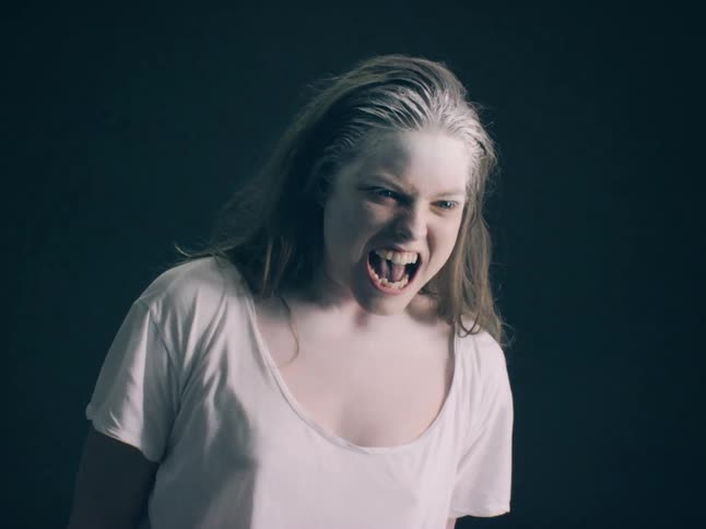 A close view shows a person with long hair and white powder on their face shouting while looking toward the camera against a dark background