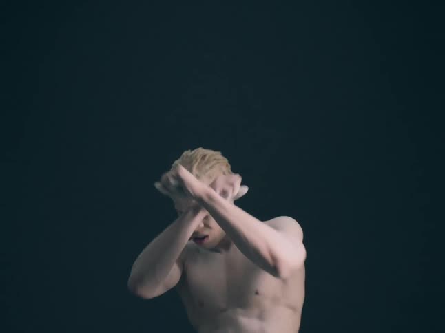 A shirtless man with skin and hair covered in white powder performs fluid and expressive movements with his arms and torso against a solid dark background in a dim studio