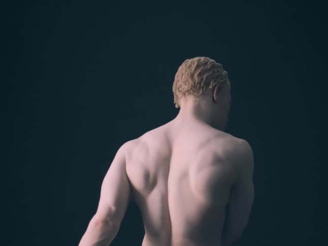 A view from behind shows the muscular back and shoulders of a shirtless man with short blonde hair standing still against a solid dark background in a dim studio