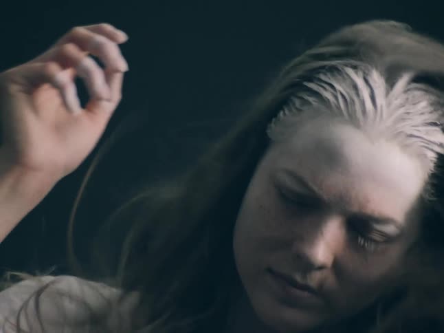 In this slowmotion clip a woman is seen with white powder possibly flour or theatrical dust scattered across her hair and forehead she moves her hands near her face as the particles drift through the air against a dark moody background
