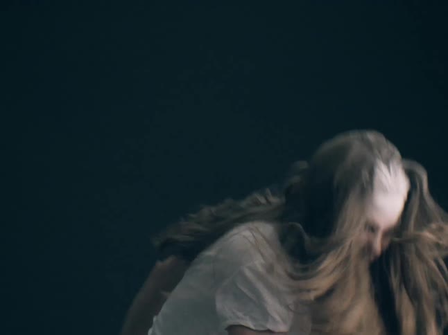 A woman wearing a white shirt with white powder on her forehead and hair moves her head and body energetically against a dark background in a dim studio