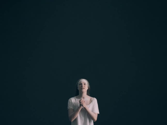 A woman stands against a dark background with her hands clasped together looking upward as if in prayer or plea over the course of the clip her expression shifts from hopeful anticipation to an openmouthed cry or shout
