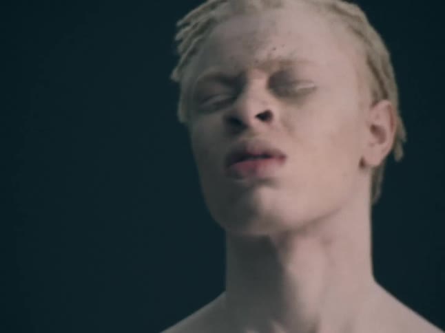 A shirtless man with albinism moves and breathes against a dark studio background