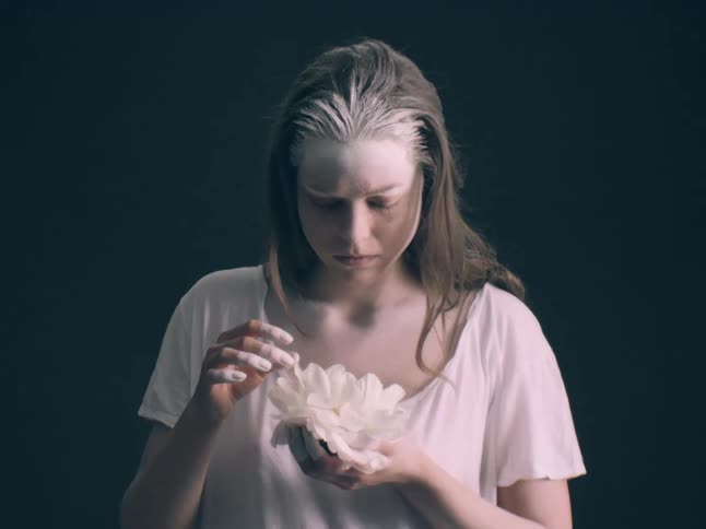 A close view shows a person with long hair and white powder on their forehead looking down at a large white flower held in their hands against a dark background